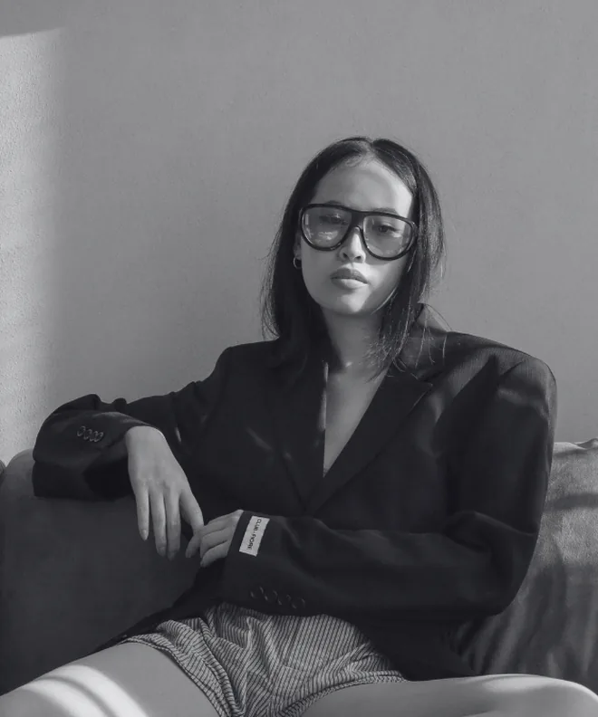 A Deely Creators model seated on a sofa in an oversized blazer and oversized glasses.